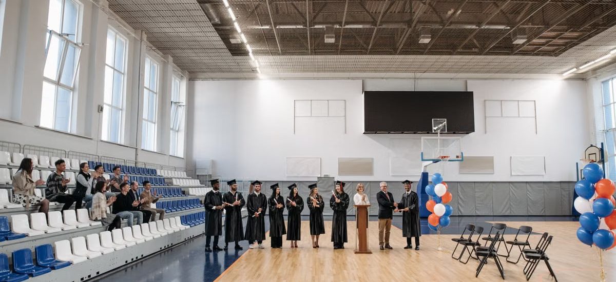 A graduation ceremony held in an indoor gymnasium with graduates and an audience.
